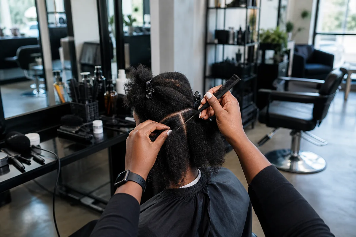 Coiffeuse traçant une raie fine au peigne queue pour poser des nattes collées sur cheveux crépus